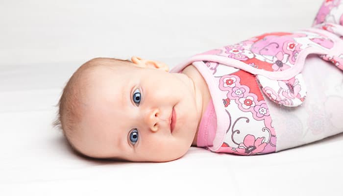 Three month baby girl wrapped in a modern winged baby swaddle laying on a bed